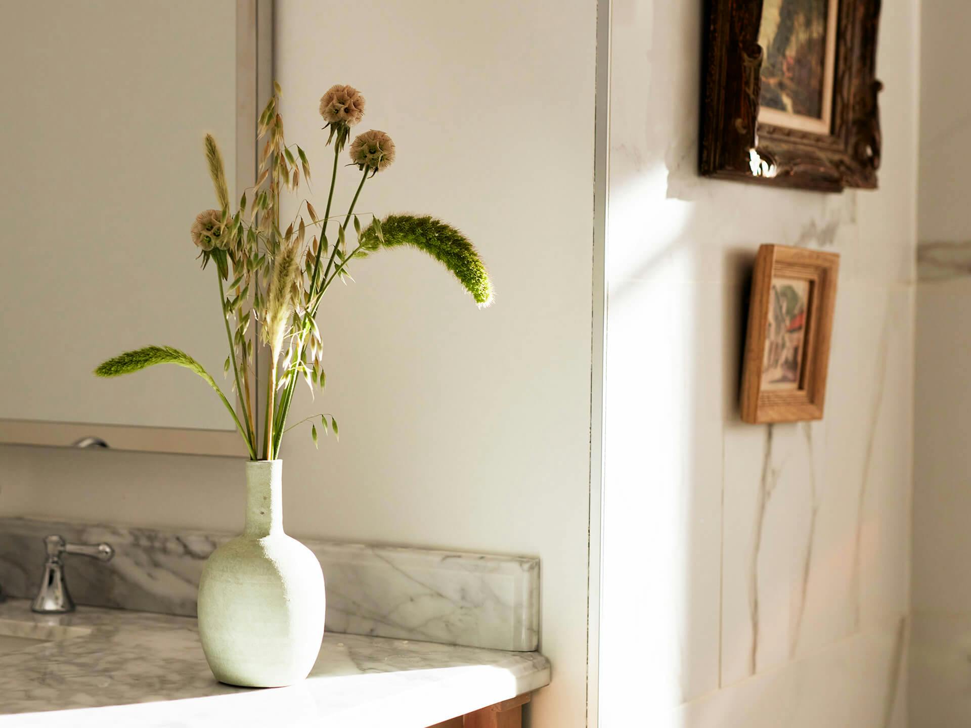 Close up of a bathroom counter with a vase of wild greenery and two small antique framed images on a wall
