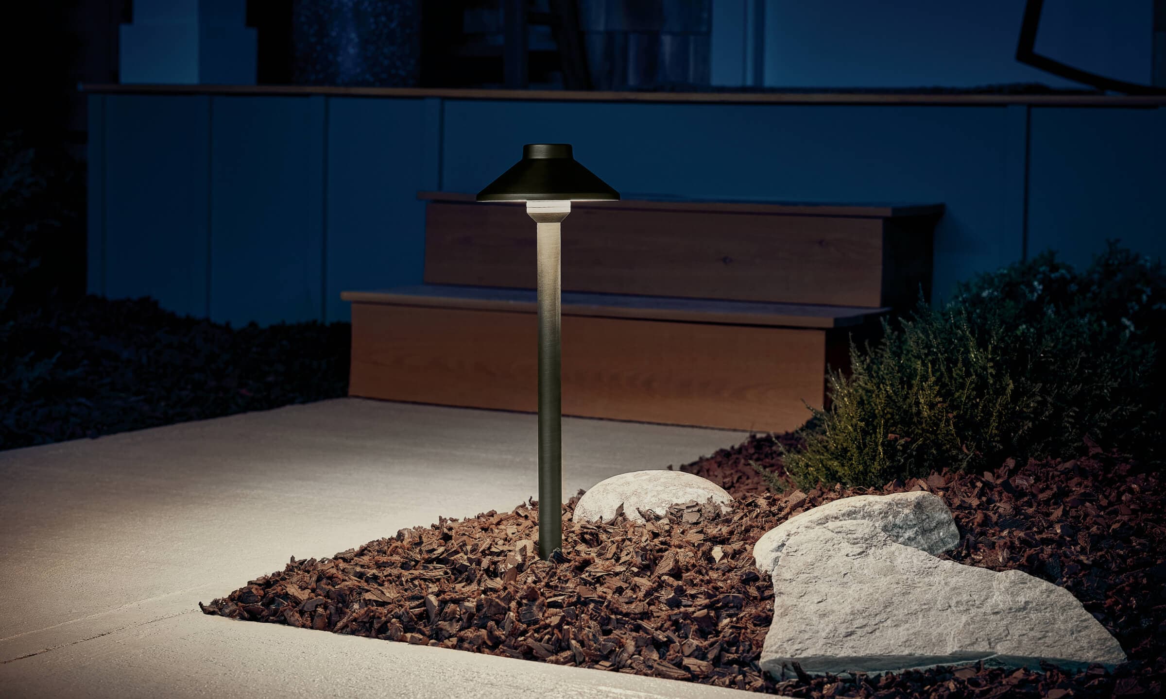 Detail shot of a Stepped Dome Path Light in landscape with mulch lighting a sidewalk turned on at night 